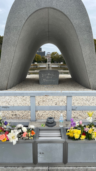 You can look through the arch to a burning flame, then beyond that to a building that still shows the marks of the atomic bomb.